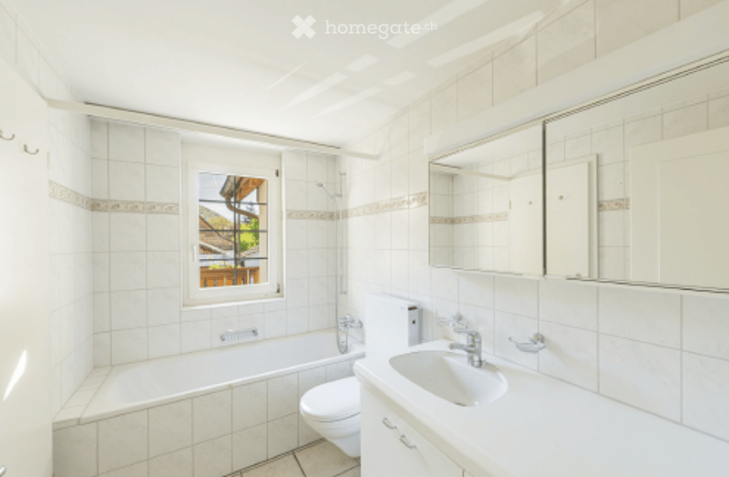 Bright bathroom with white tiles, a bathtub, toilet, sink, and a large mirror above the sink. A window offers a view of the outdoor area.