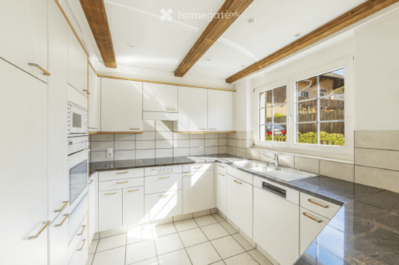 Bright, modern kitchen with white cabinets, stainless steel appliances, and exposed wooden beams on the ceiling. Features a large window with a view of the garden.