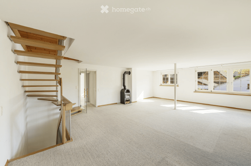 Empty room with white walls, wooden staircase, fireplace, and large windows.