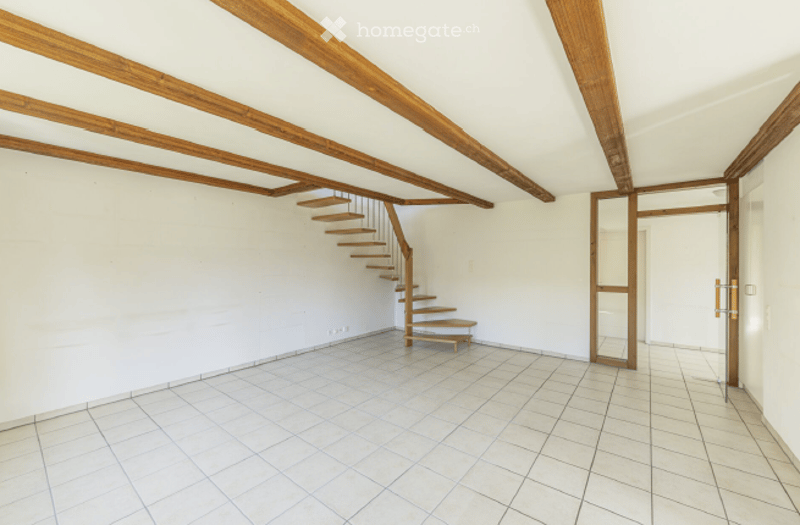 Empty room with white walls, exposed wooden beams, tiled floor, and a small wooden staircase leading to an upper level.