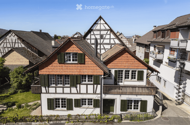 Traditional half-timbered house with red-tiled roof, green shutters, and white walls, surrounded by neighboring houses and greenery.