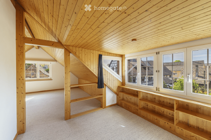 Bright, airy attic space with wooden beams, built-in shelving, large windows, and a skylight. No furniture present.