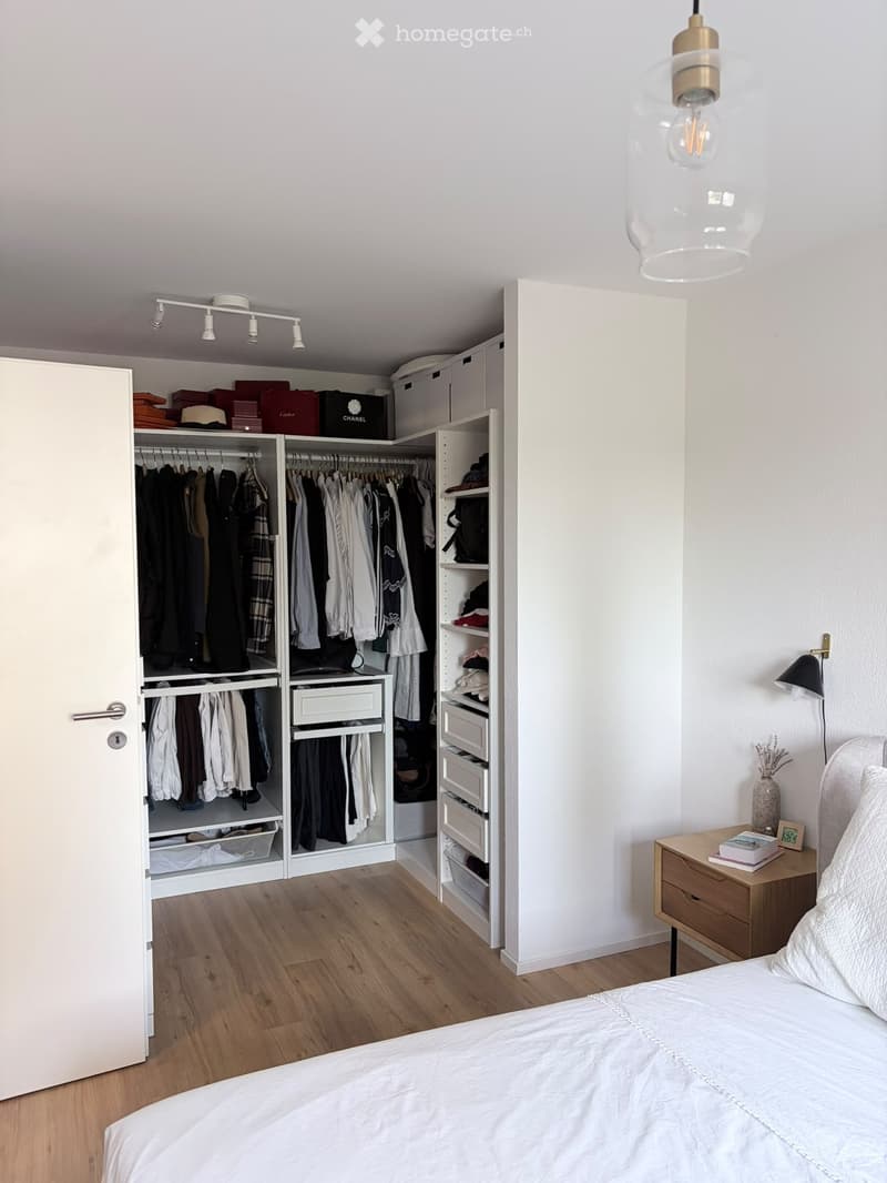 Modern bedroom with a large bed, nightstand, wardrobe with hanging clothes, and a pendant light fixture.