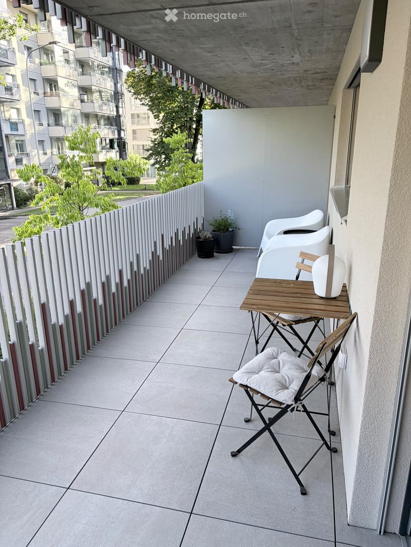 Balcony with a small wooden table, white chairs, and potted plants, overlooking a cityscape with trees and buildings.