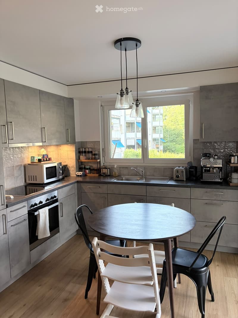 Modern kitchen with grey cabinets, wooden flooring, round dining table with chairs, microwave, oven, sink, toaster, coffee machine, and pendant lights.