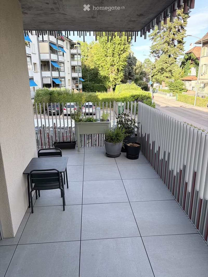 Balcony with tiled floor, metal table and chairs, potted plants, and a view of the street and surrounding buildings.