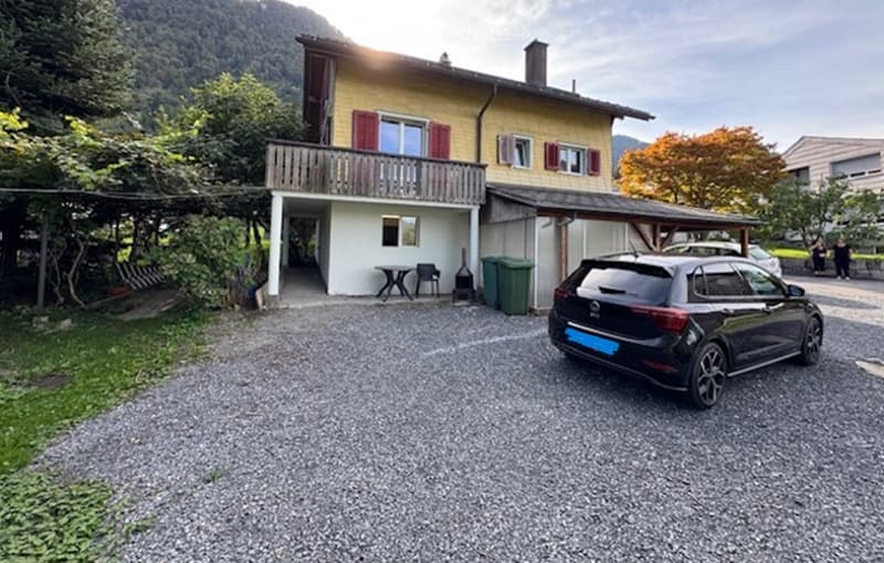 Two-story house with yellow facade, red shutters, balcony, and parking area with a car.
