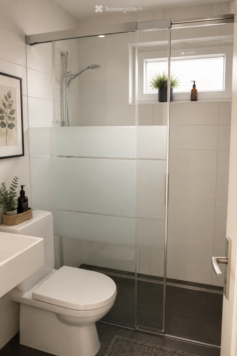 Modern bathroom with white tiles, a glass-enclosed shower, a toilet, a sink, and a framed picture on the wall. There are small plants and bottles on the windowsill and sink counter.