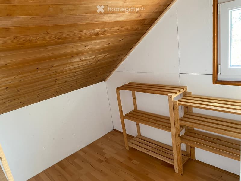 Empty room with wooden floor, white walls, and a wooden shelf unit against the wall. The ceiling has wooden paneling with a sloped design.