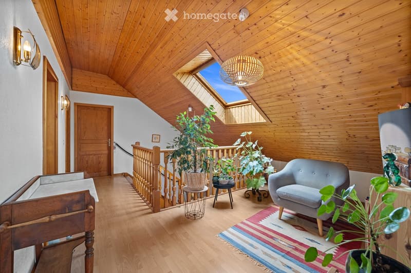 Cozy, wooden-paneled room with a slanted ceiling, skylight, and a staircase leading to an upper level. Features a grey sofa, small round table, potted plants, and a colorful rug.