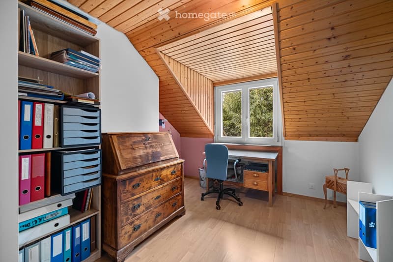 A cozy attic study with wooden beams, a large window, a wooden desk, a blue office chair, a bookshelf filled with books and folders, and a small wooden cabinet.