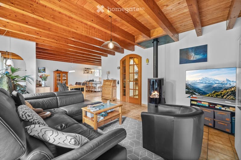 Living room with exposed wooden beams, a black leather sofa, a coffee table, an armchair, a fireplace, a large TV, and a dining area with a table and chairs.