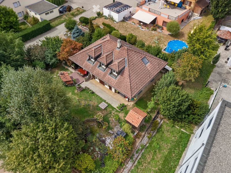 Aerial view of a two-story house with a red-tiled roof, surrounded by a well-maintained garden, patio, and outdoor seating area. The property includes a swimming pool, swing set, and various trees and shrubs.