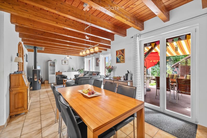 Bright living and dining area with exposed wooden beams, a large dining table with chairs, a sofa, an armchair, a fireplace, and a view to a patio.