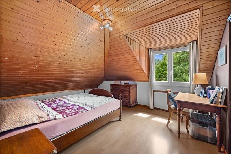 Cozy bedroom with wooden walls and ceiling, featuring a bed with patterned bedding, a nightstand, a wooden desk with a chair, and a window with curtains.