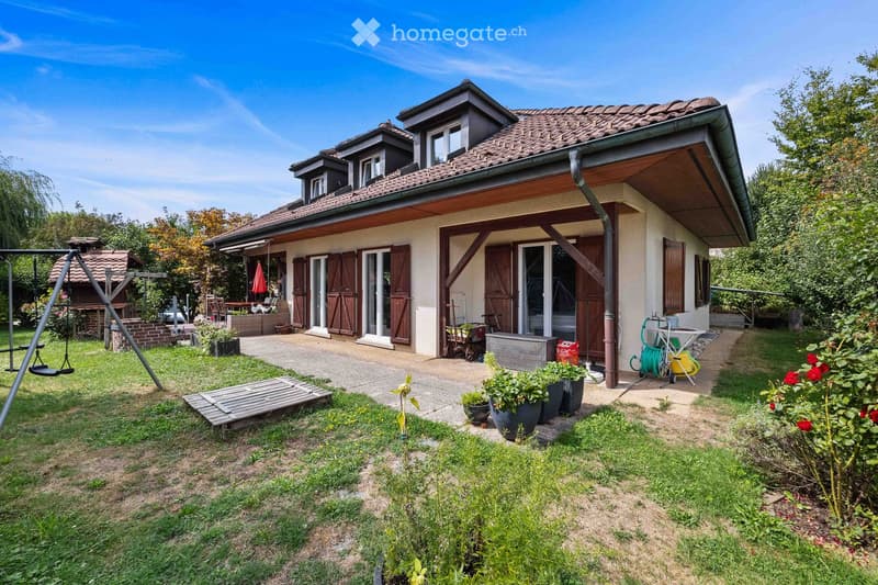 Two-story house with a tiled roof, wooden shutters, and a covered entrance. The garden includes a swing, potted plants, and a small patio area.