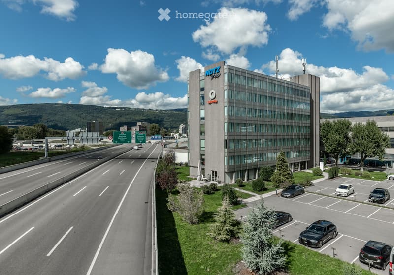 A multi-story office building with the Notz logo on the facade, surrounded by a parking lot and a highway with multiple lanes of traffic.