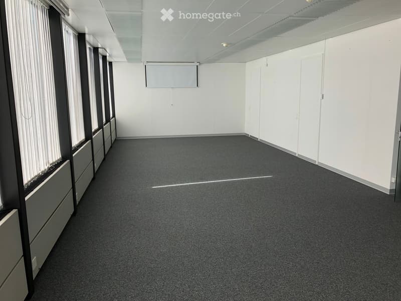 Large open room with gray carpeted floor, white walls, and a projector screen mounted on the wall. The room appears to be an office or meeting space with ample natural light from the windows.