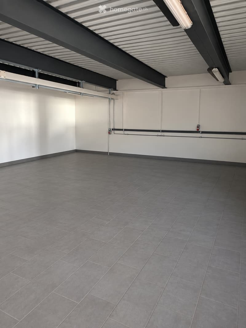 Empty room with grey tiled floor and white walls, featuring exposed metal beams and lighting fixtures on the ceiling.