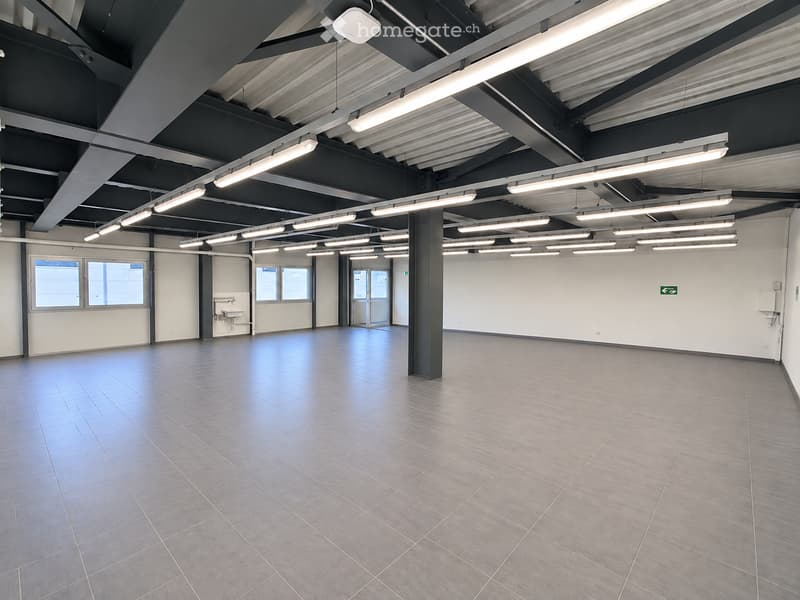 Empty room with tiled floor, white walls, exposed ceiling beams, and fluorescent lighting. Features include a sink, windows, and a central pillar.
