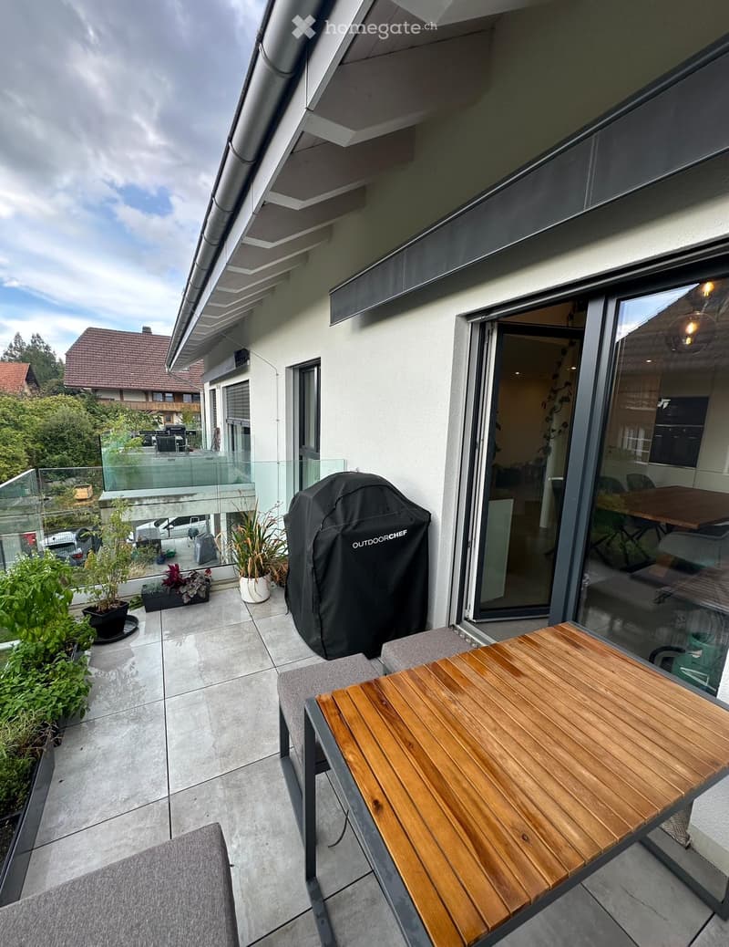 Balcony with a wooden table, grey chair, potted plants, and a covered grill. The balcony has a modern glass railing and overlooks a residential area.