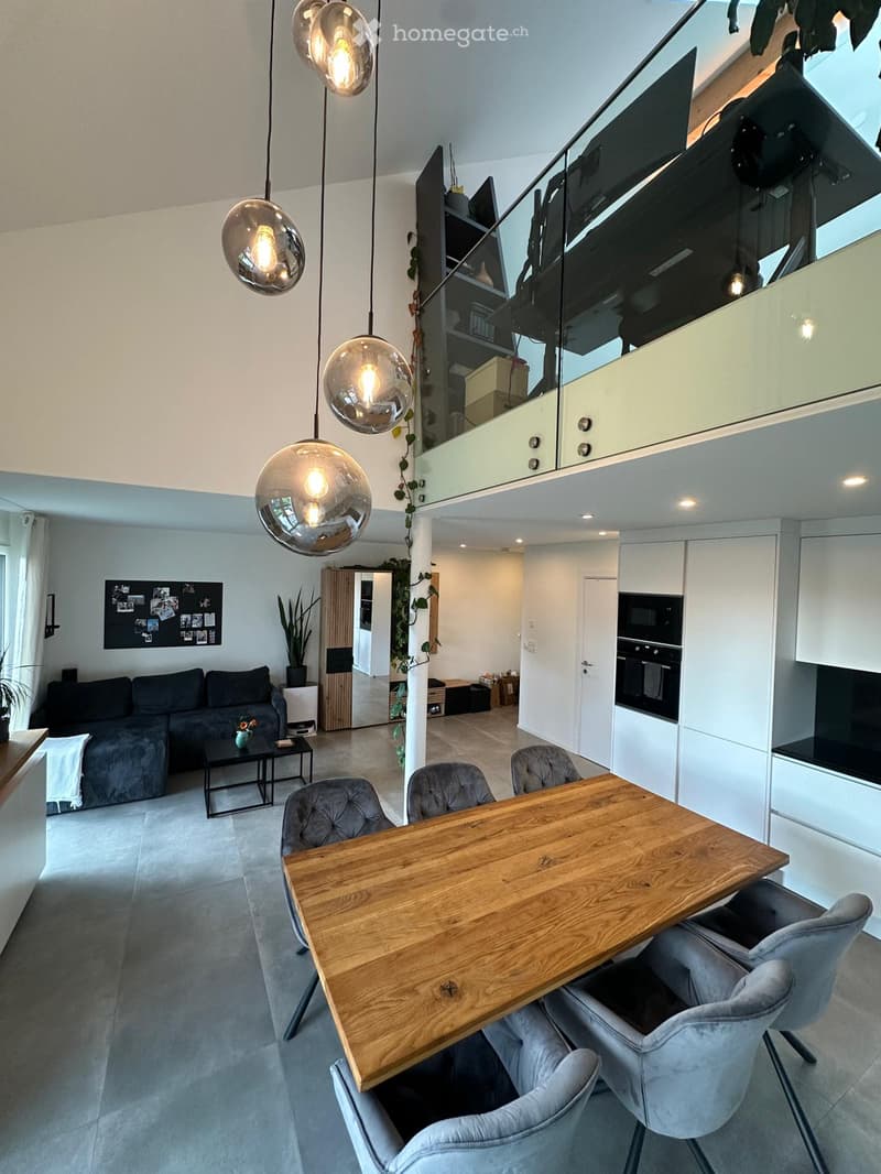 Modern open-plan living and dining area with grey sofa, coffee table, wooden dining table, grey chairs, pendant lights, and white cabinets.