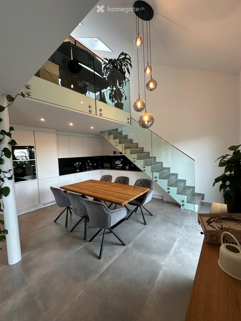 Modern open-plan kitchen and dining area with grey chairs, wooden dining table, white cabinets, and a sleek staircase leading to an upper level.