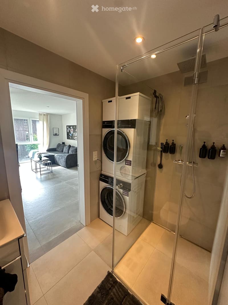 Modern bathroom with a glass-enclosed shower, a washing machine and dryer unit, and a towel on the floor. The room has tiled flooring and walls, with a view into an adjacent living space.