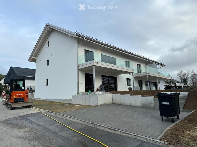 Modern two-story house with white exterior, large windows, and a raised ground floor. Features include a balcony, garage, and construction equipment nearby.
