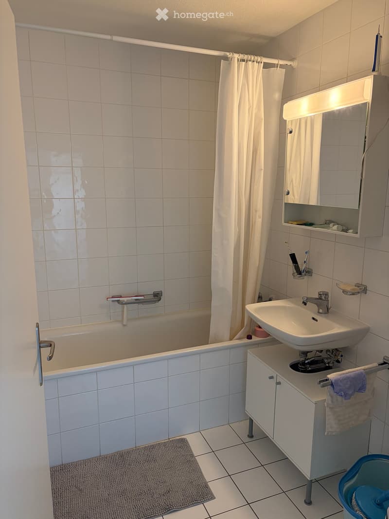 A small bathroom with a bathtub, a sink with a mirror cabinet, a towel on the cabinet, and a toothbrush holder on the wall.