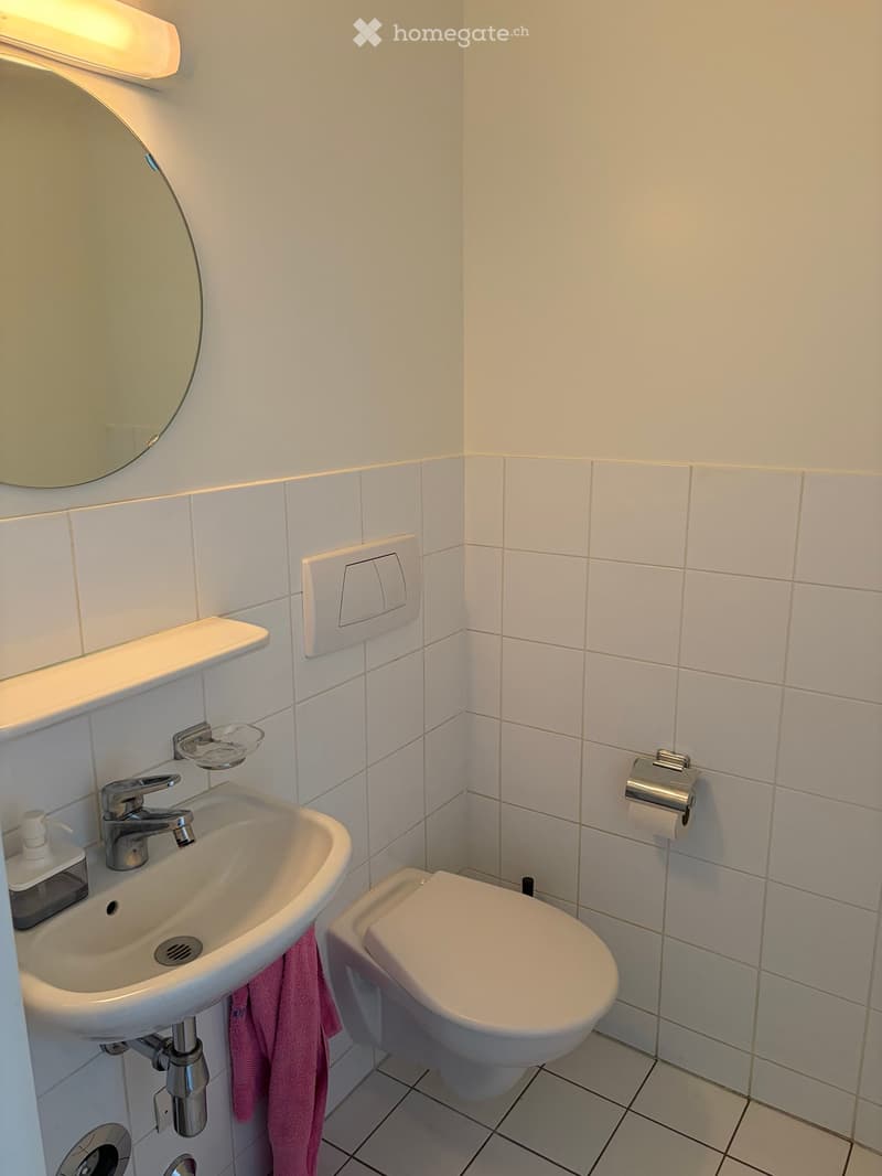Modern bathroom with white tiles, a sink with a modern faucet, a round mirror, a toilet, and a towel hanging on the side of the sink.