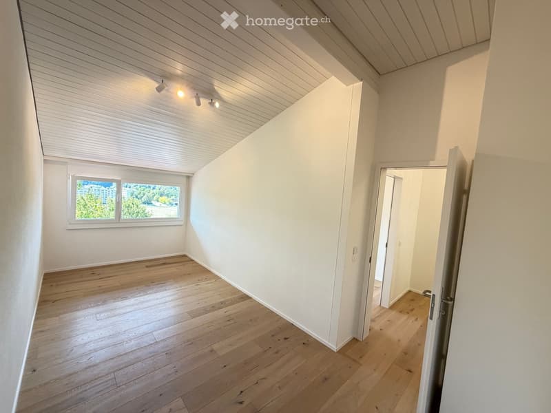Empty room with wooden flooring, white walls, and a large window with blinds. The ceiling has recessed lighting and a slanted design, typical of an attic or loft space.
