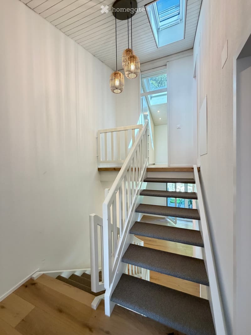 Modern staircase with wooden steps, white railing, and pendant lights, leading to a bright, open area with skylights.