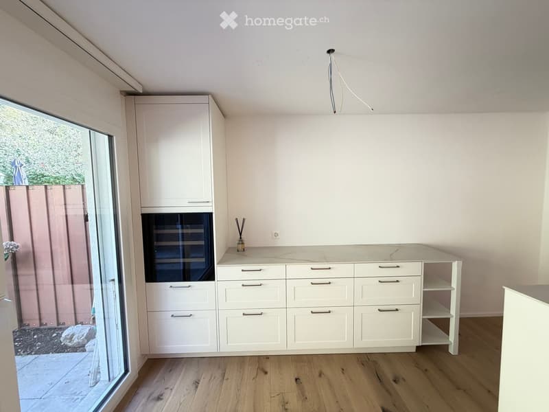 Modern kitchen with white cabinets, a built-in oven, and a marble countertop. The space features wooden flooring and large windows with a view of the garden.
