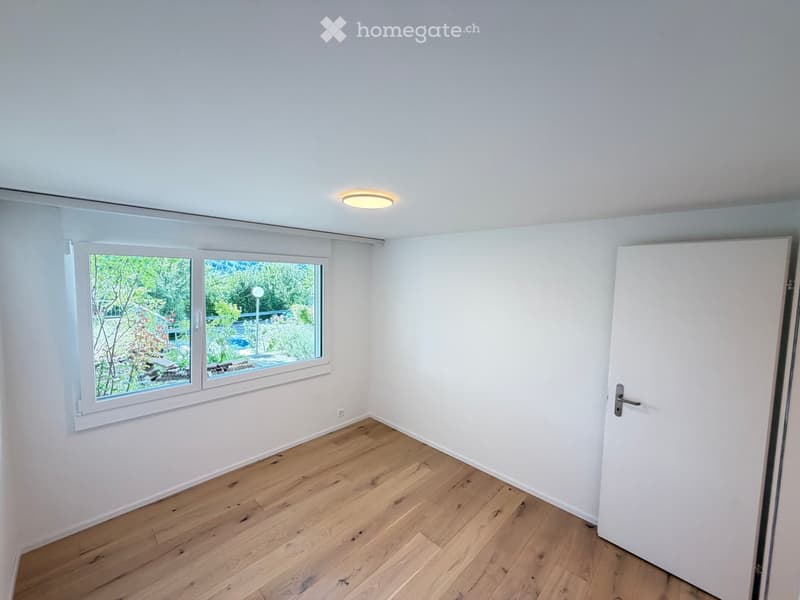 Empty room with light wooden flooring, white walls, large window with view of greenery, and a closed white door.