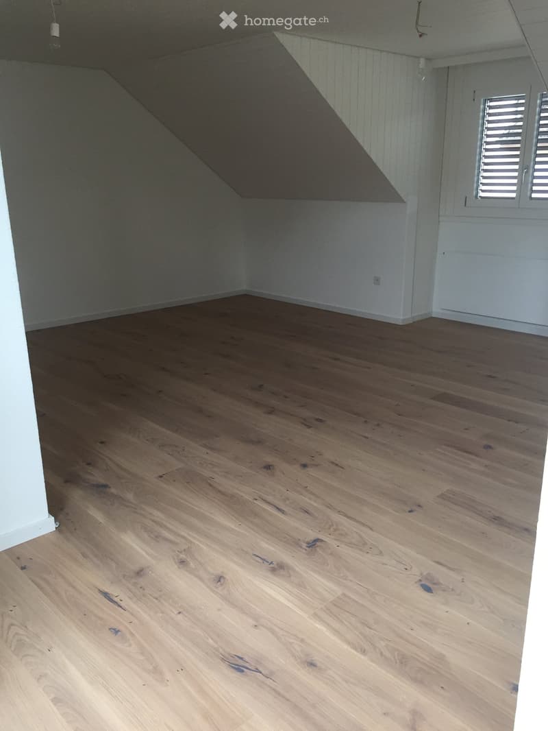 Empty room with wooden flooring and white walls, featuring a slanted ceiling and a window with shutters.