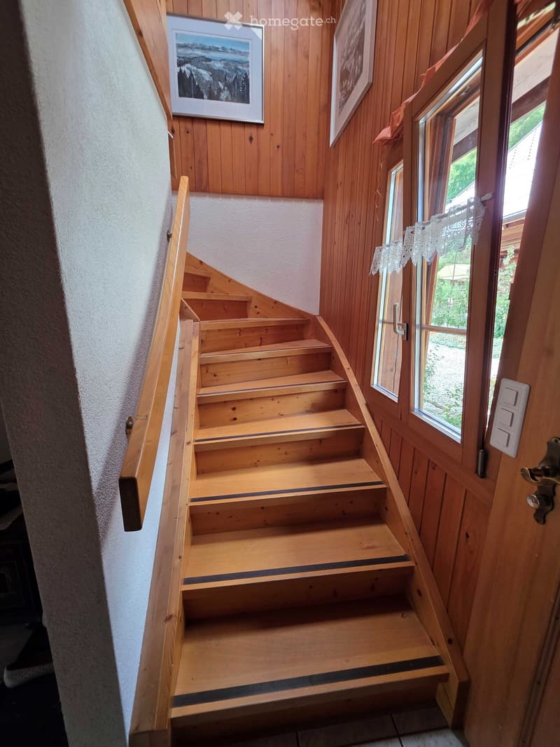 Wooden staircase with handrail, white wall, framed pictures, and a window with lace curtains.