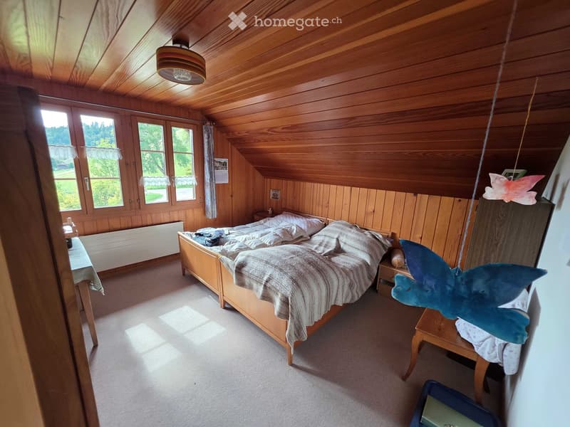Cozy attic bedroom with wooden walls and ceiling, large windows, a double bed with pillows and blankets, nightstands, and decorative items.
