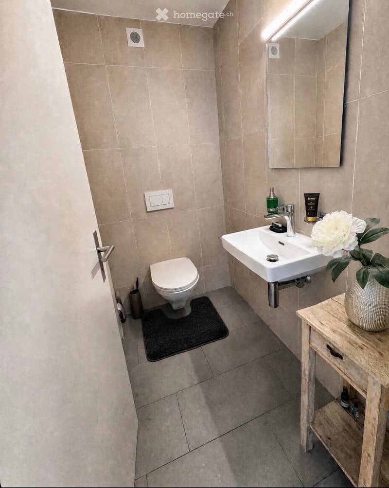 Modern bathroom with beige tiles, a toilet, a sink with a modern faucet, a mirror, a small wooden table with a vase of white flowers, and a black bath mat.