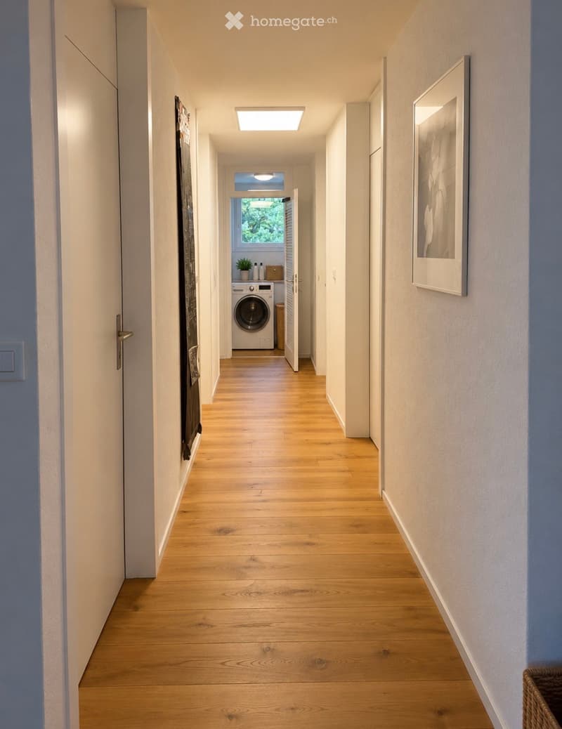 Modern hallway with light wooden flooring, white walls, and recessed ceiling lighting. A framed picture hangs on the right wall, and a small laundry area is visible at the end.