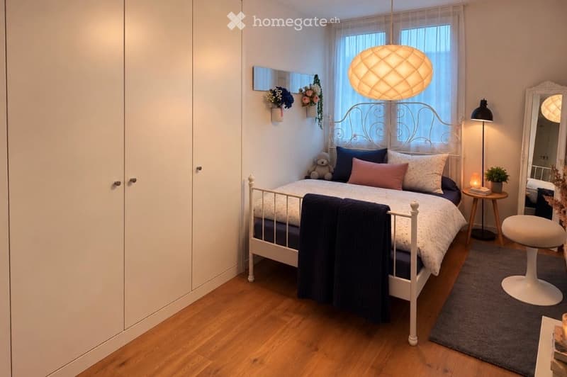 Cozy bedroom with a white wooden bed, multiple pillows, a blue blanket, a small round table with a lamp, a white stool, a large mirror, and a small plant on the table.