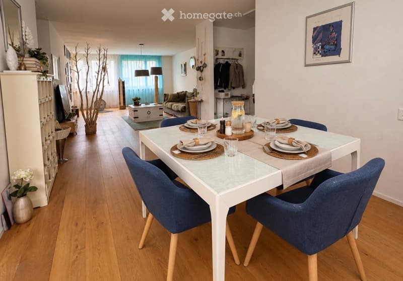 Open-plan living and dining area with wooden flooring, white dining table with blue chairs, sofa, lamps, and decorative items.