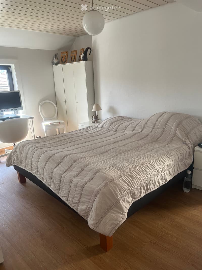A modern bedroom with a large bed, a white chair, a small table with a lamp, a cabinet, and a computer screen on a desk.