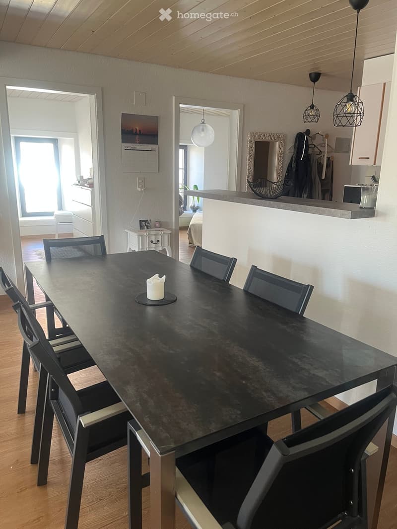Dining area with a black dining table surrounded by black chairs, a white candle in the center, and a modern interior with wooden flooring and pendant lights visible in the background.