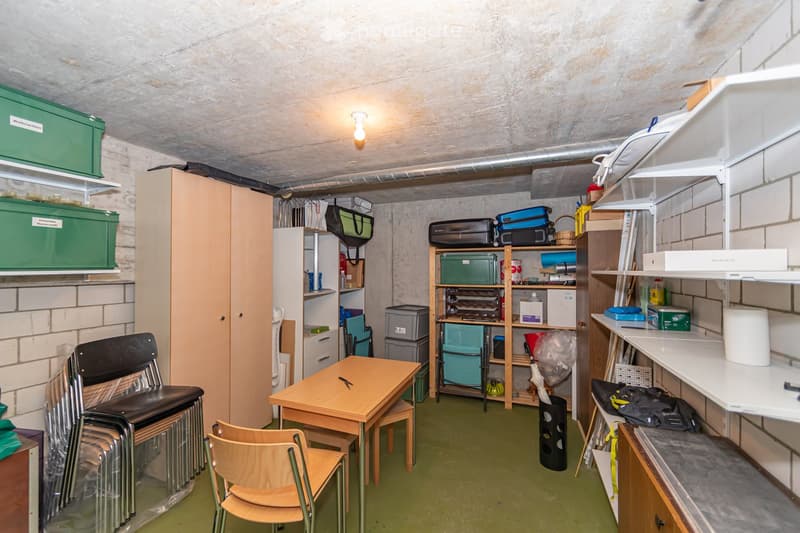 A small, furnished room with a table, chairs, shelves, storage boxes, and a laptop. The room has exposed brick walls and a concrete ceiling.