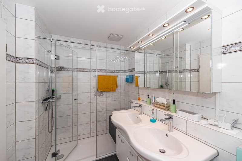 Modern bathroom with white marble tiles, a glass-enclosed shower, double sink vanity, and various toiletries.