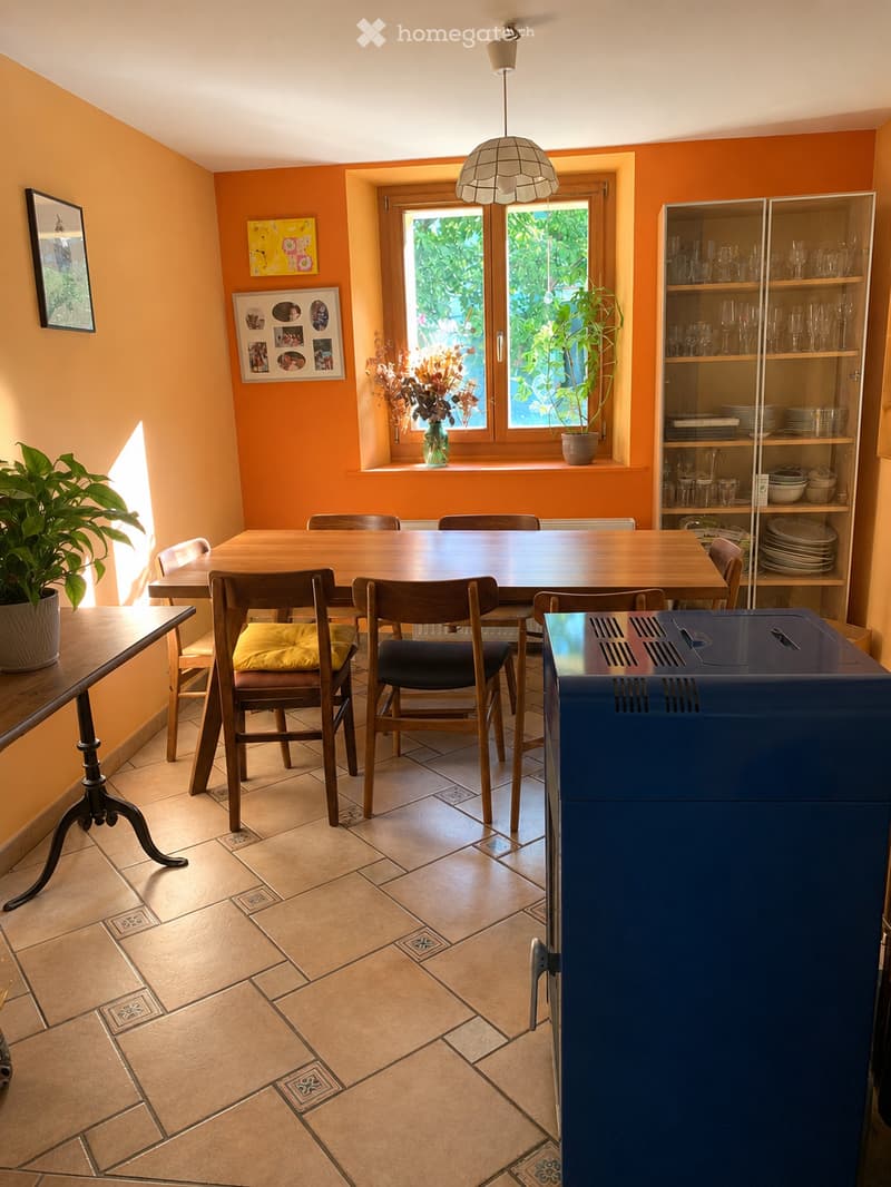 Bright dining area with a wooden table, chairs, a glass cabinet, and potted plants. The room features warm orange walls, tiled flooring, and large windows with a view of greenery.