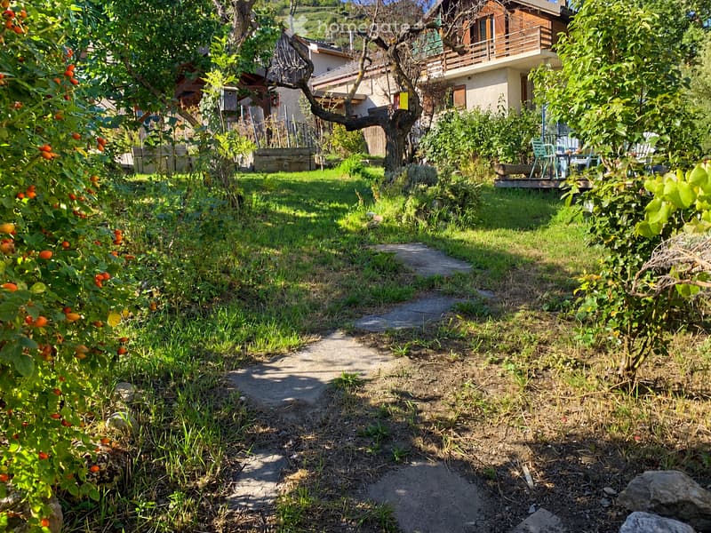 A lush garden with a pathway leading to a house, featuring fruit trees, grass, and outdoor seating on a wooden deck.