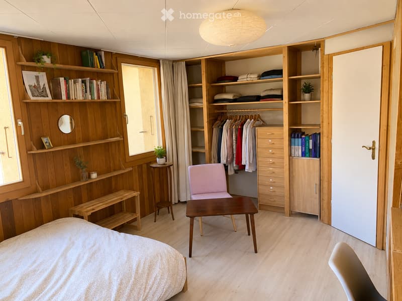 Cozy bedroom with wooden walls and flooring, featuring a bed, a small table, a pink chair, a bench, a built-in wardrobe with hanging clothes, and shelves with books and plants.