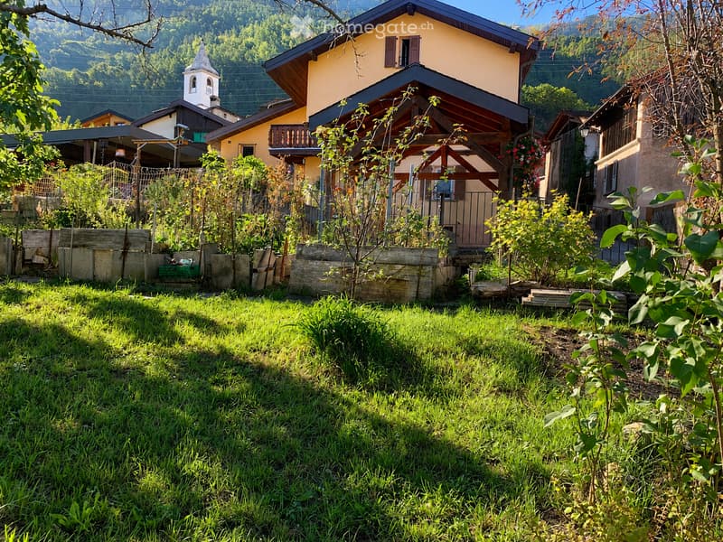 A well-maintained garden with a grassy lawn, wooden planters, and a small wooden fence. The background features a yellow house with a balcony and a small chapel in the distance.